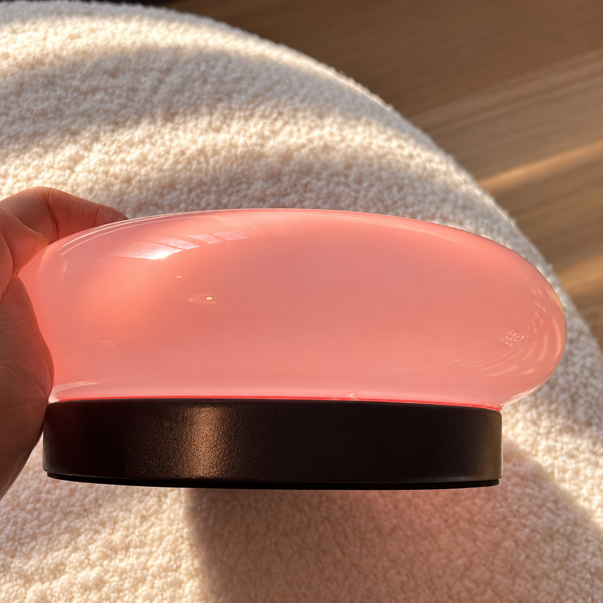 Side view of a pink glass donut lamp held above cream textured fabric, showing its dark circular base.