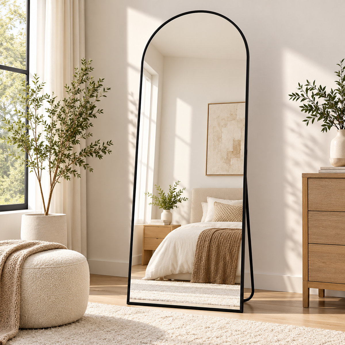 Black frame arched standing mirror reflecting a bed, positioned beside a window, potted tree, ottoman, and dresser by HavvakHome.