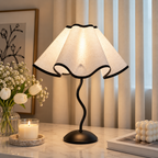 Scalloped cream table lamp with a wavy black stem beside white tulips, a lit candle and stacked books.