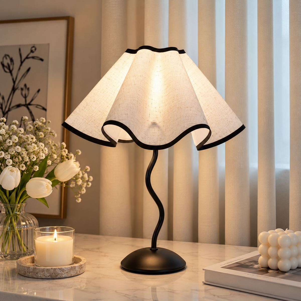 Scalloped cream table lamp with a wavy black stem beside white tulips, a lit candle and stacked books.