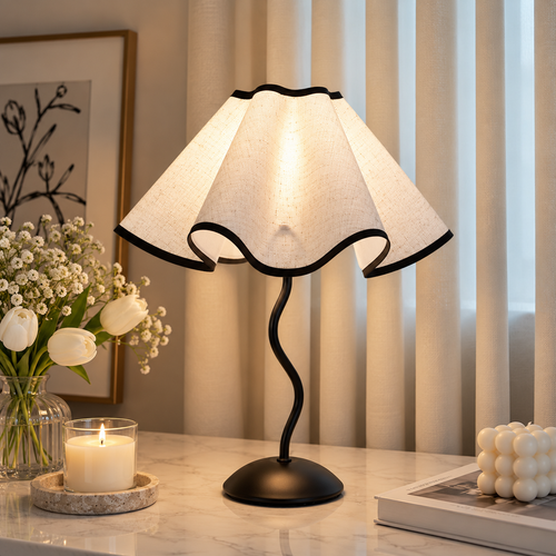Scalloped cream table lamp with a wavy black stem beside white tulips, a lit candle and stacked books.