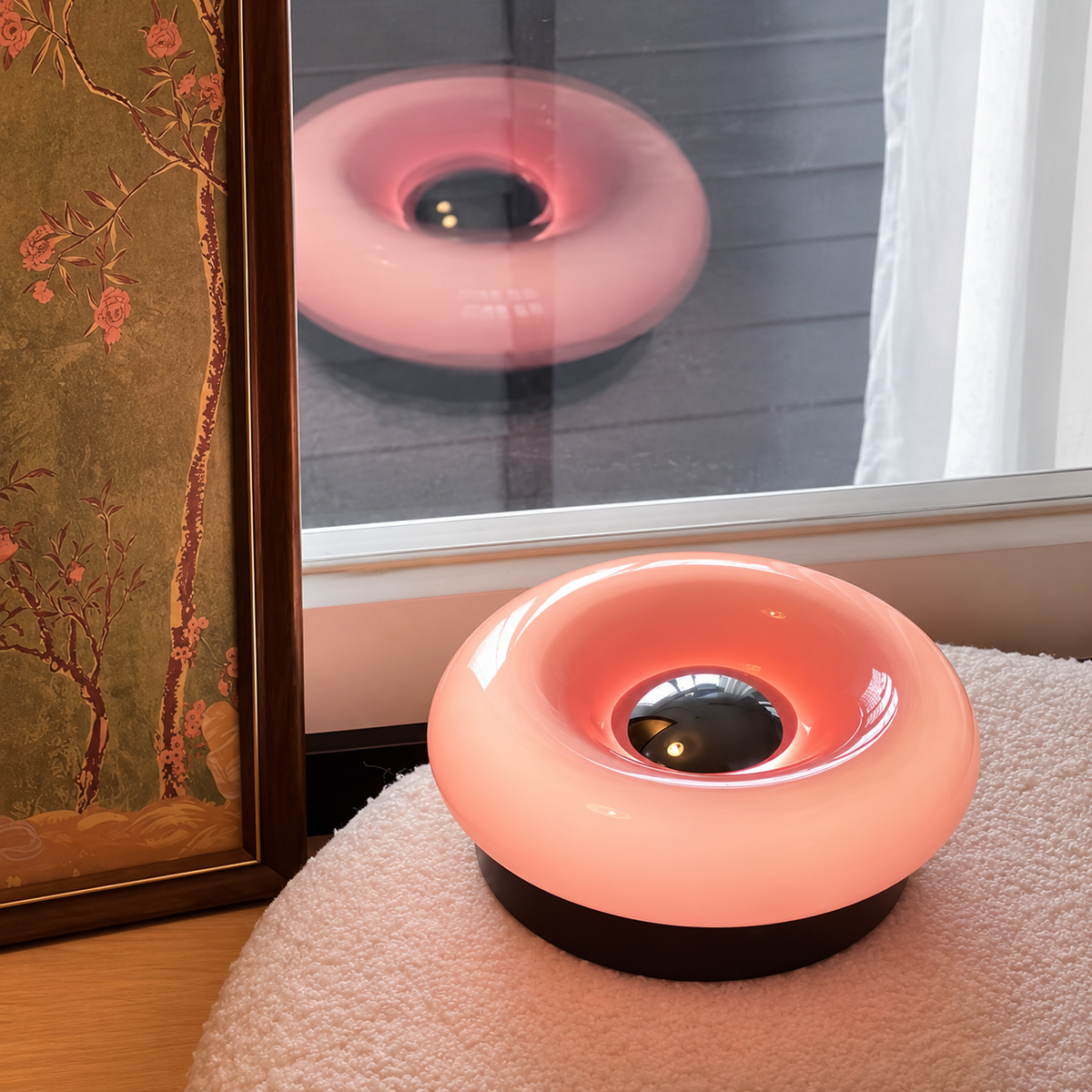 Pink donut table lamp with a dark circular base on textured fabric beside a window and framed floral artwork.