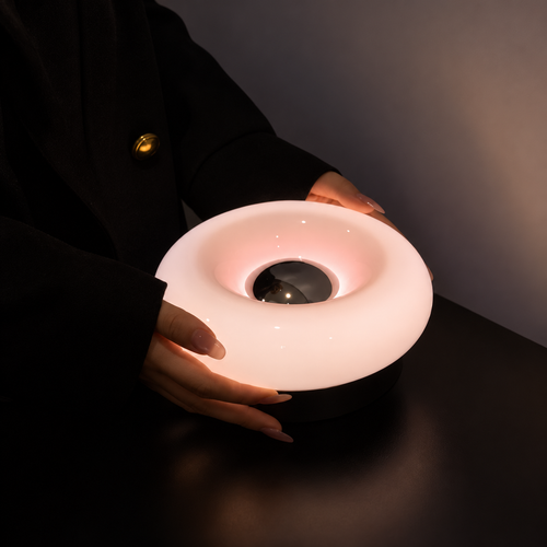 Hands holding a glowing pale pink donut-shaped lamp in a dimly lit room.