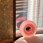 Pink donut-shaped lamp standing upright beside a sunlit window, with a reflective center and floral artwork nearby.