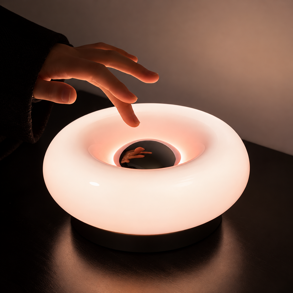 Hand reaching toward the reflective center of an illuminated pink donut lamp on a dark table.