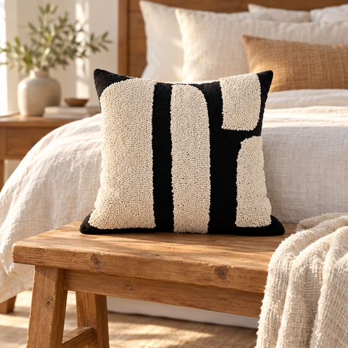 Square black and cream geometric tufted pillow cover on a wooden bench at the foot of a linen bed.