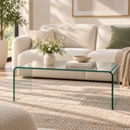 Clear bent-glass waterfall coffee table holding a white flower vase, books, and a bowl in front of a cream sofa with a sage pillow in a sunny living room by HavvakHome.