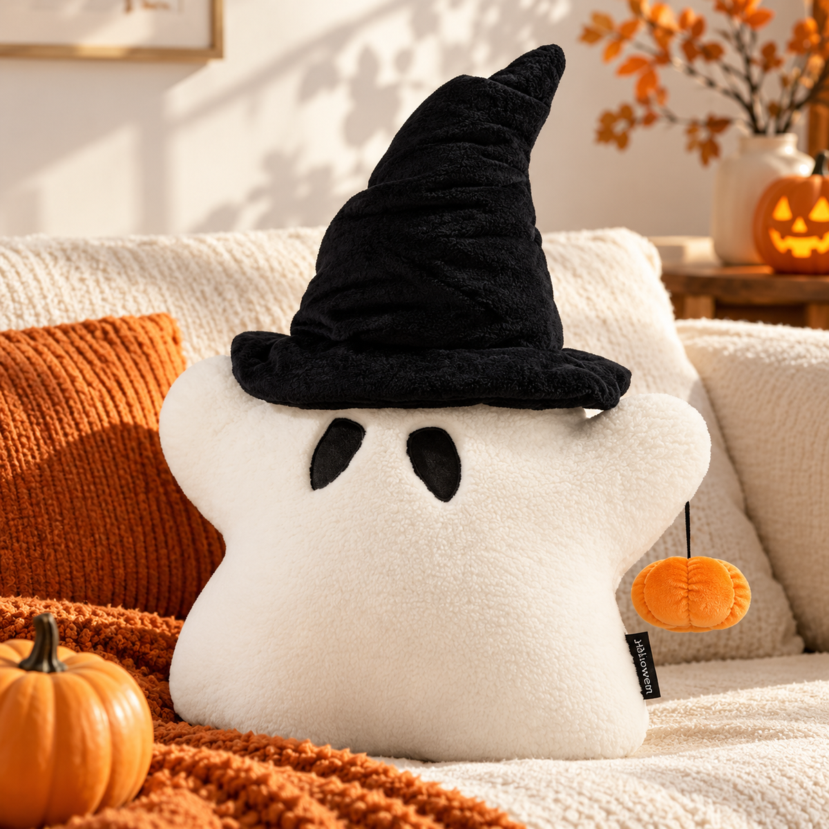 Off-white ghost pillow wearing a black witch hat with a dangling orange pumpkin displayed on a cream sofa with an orange cushion and blanket by HavvakHome.