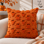 Single orange raised ghost-pattern pillow on a cream sofa styled with a knitted throw and Halloween pumpkins in the background.
