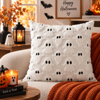 Single white raised ghost-pattern pillow with black eyes on a cream sofa styled with an orange knitted blanket and Halloween decorations.