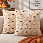 Two cream raised ghost-pattern pillows with black eyes on a cream sofa styled with an orange knitted throw and pumpkins.
