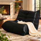 Black button-tufted armless floor lounge chair with a cream knitted throw on a rug beside a fireplace and round wood side table by HavvakHome.