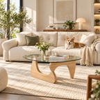 Clear triangular glass coffee table with a light wood sculptural base, flower vase, books, and bowl before a cream sofa with a corgi in a sunny living room.