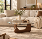 Walnut-toned sculptural wood base and rounded triangular clear glass top styled with a greenery vase, books, candle, and wooden bowl before a cream sectional sofa.