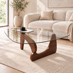 Rounded triangular clear-glass coffee table with a sculptural walnut-tone wood base, styled with a black cup and book before a cream sofa.