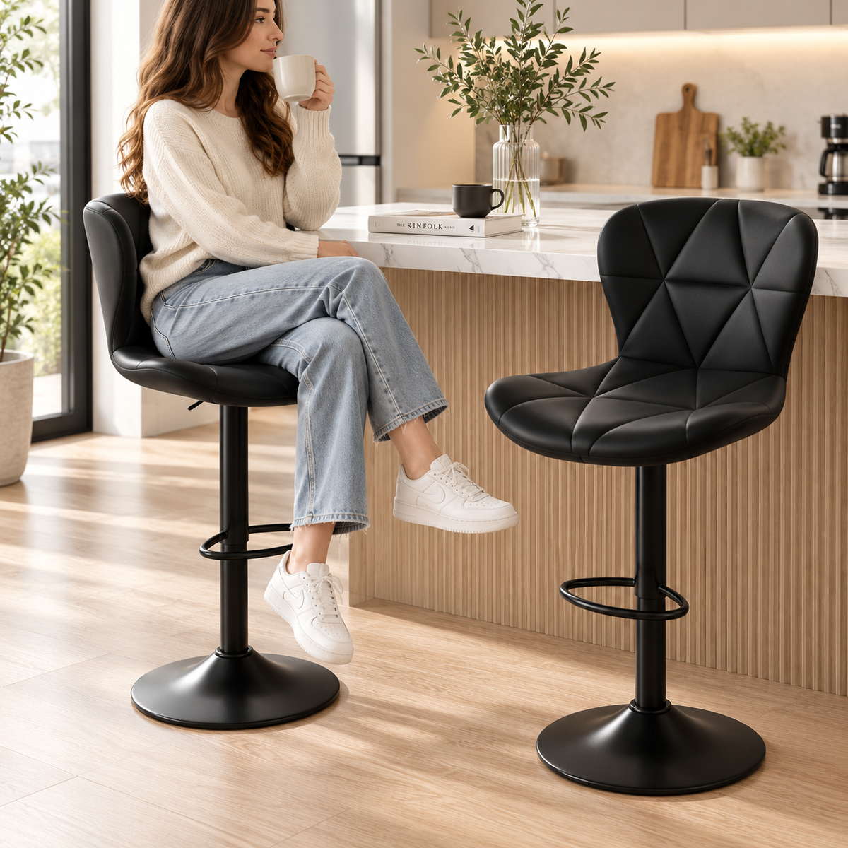 Woman holding a mug seated on a black quilted bar stool beside a second black bar stool with black pedestal bases and footrests at a kitchen island by HavvakHome.