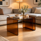 Amber tinted bent glass table placed in front of a gray sectional sofa, styled with books, a bowl, flower vase, and candle by HavvakHome.
