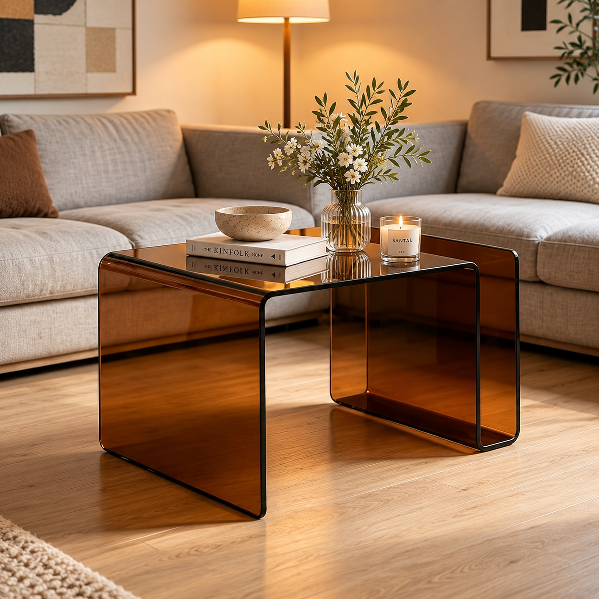 Amber tinted bent glass table placed in front of a gray sectional sofa, styled with books, a bowl, flower vase, and candle by HavvakHome.