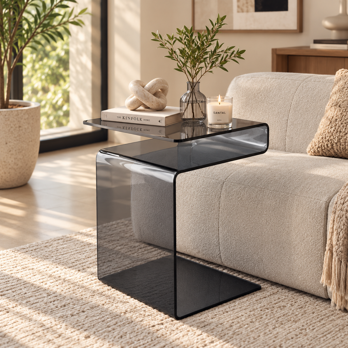 Smoke gray S-shaped glass side table beside a cream sofa styled with a knot ornament, book, candle, and leafy vase by HavvakHome.