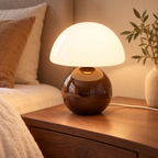 Coffee ceramic mushroom lamp with a glossy rounded base and a white glass dome shade on wooden bedside furniture in a softly lit bedroom.