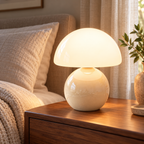 Cream ceramic mushroom lamp with a glossy rounded base and a white glass dome shade on wooden bedside furniture in a softly lit bedroom.
