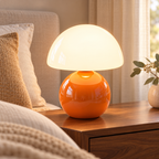Orange ceramic mushroom lamp with a glossy rounded base and a white glass dome shade on wooden bedside furniture in a softly lit bedroom.