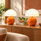 Two orange ceramic mushroom lamps with white glass dome shades on wooden furniture in a naturally lit living room.