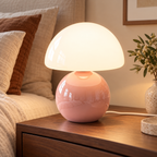 Pink ceramic mushroom lamp with a glossy rounded base and a white glass dome shade on wooden bedside furniture in a softly lit bedroom.