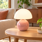 Pink ceramic mushroom lamp with a glossy rounded base and a white glass dome shade on wooden furniture in a naturally lit living room.