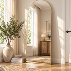 Gold arched mirror leaning beside a white door and plant, reflecting a wood cabinet and lamp by HavvakHome.
