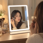Woman applying makeup with a brush reflected in an illuminated white rectangular LED travel mirror on a wooden vanity with cosmetics by HavvakHome.