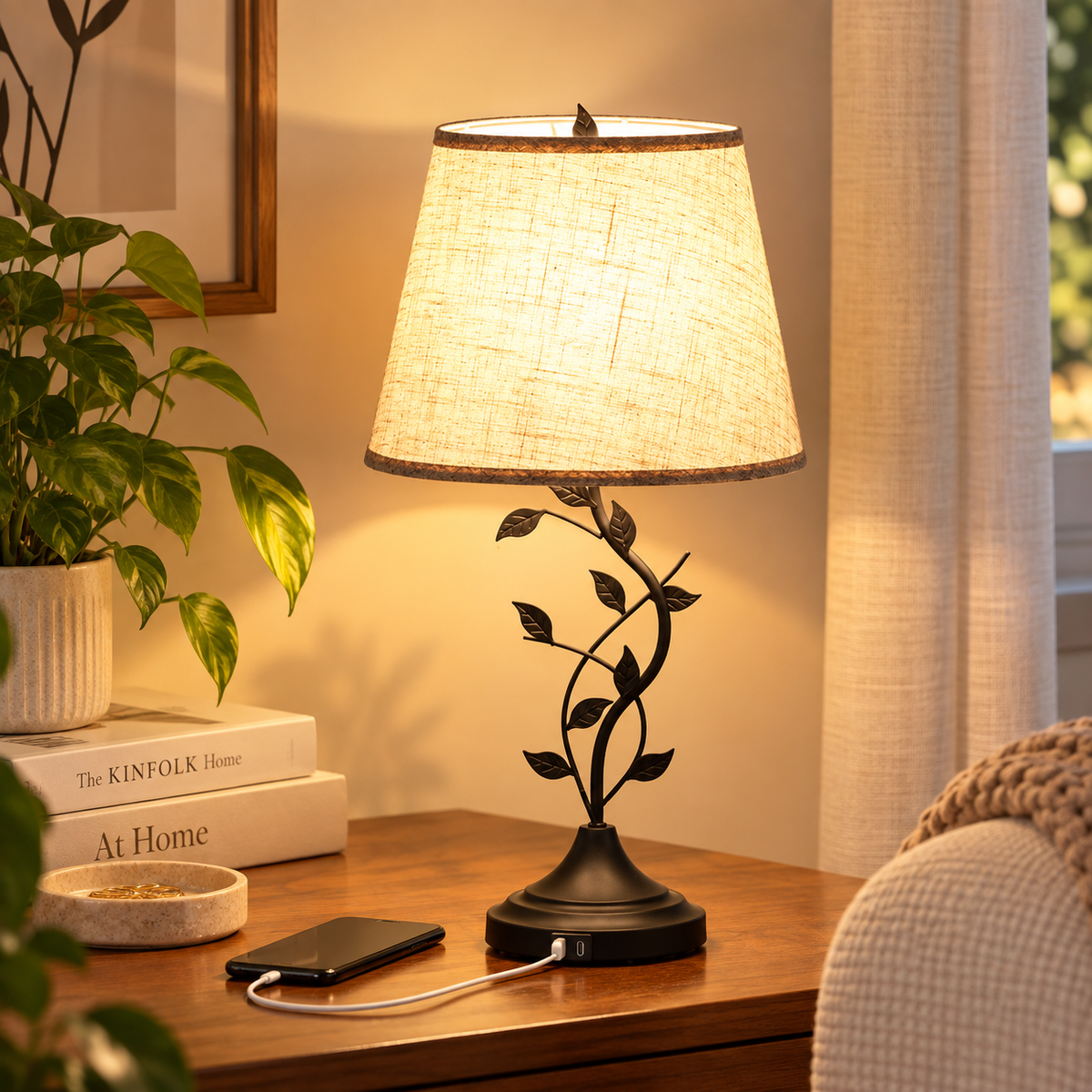 Warm glowing leaf-vine table lamp with beige tapered shade and dark base on a wood side table, with a phone charging from the base port beside books and a plant by HavvakHome.