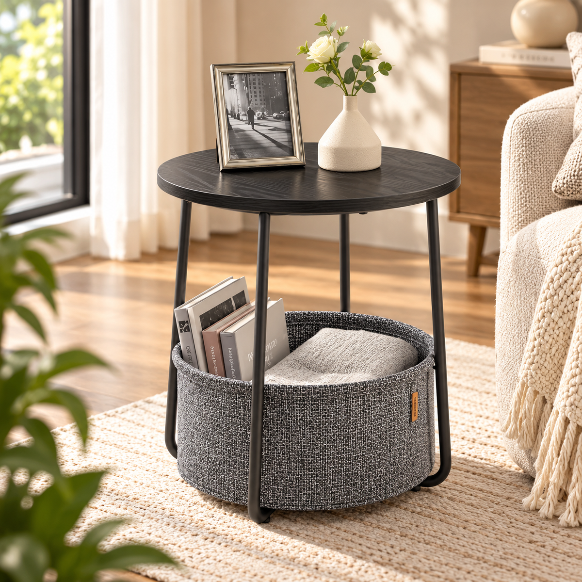 Black round side table with a dark gray basket holding books and a gray cushion beside a cream chair, with a framed photo and flowers on top by HavvakHome.