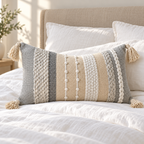 Gray 20x12 inch lumbar pillow cover with vertical woven stripes and tassels resting on a white bed with a beige headboard.