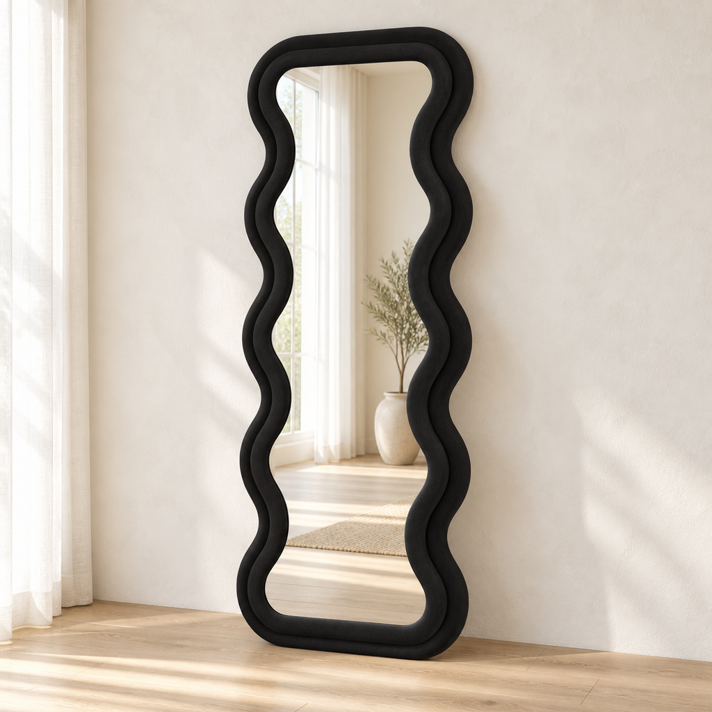 Black wavy flannel-wrapped floor mirror leaning against a pale wall, reflecting a window and vase.