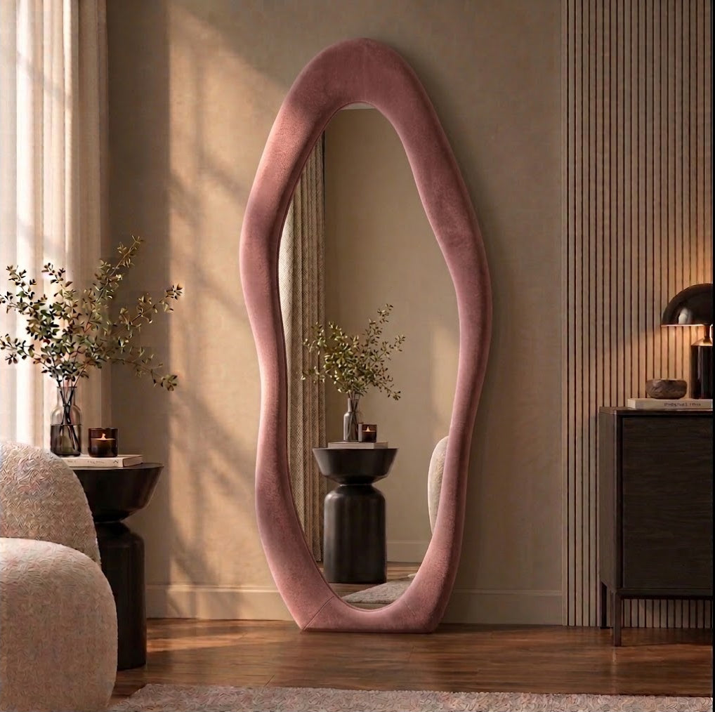 Pink wavy flannel-framed floor mirror leaning beside a wood-slat wall, reflecting a vase on a side table.