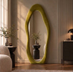 Olive green wavy flannel-framed floor mirror leaning beside a wood-slat wall, reflecting a vase on a side table.