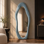 Blue-gray wavy flannel-framed floor mirror leaning beside a wood-slat wall, reflecting window curtains.