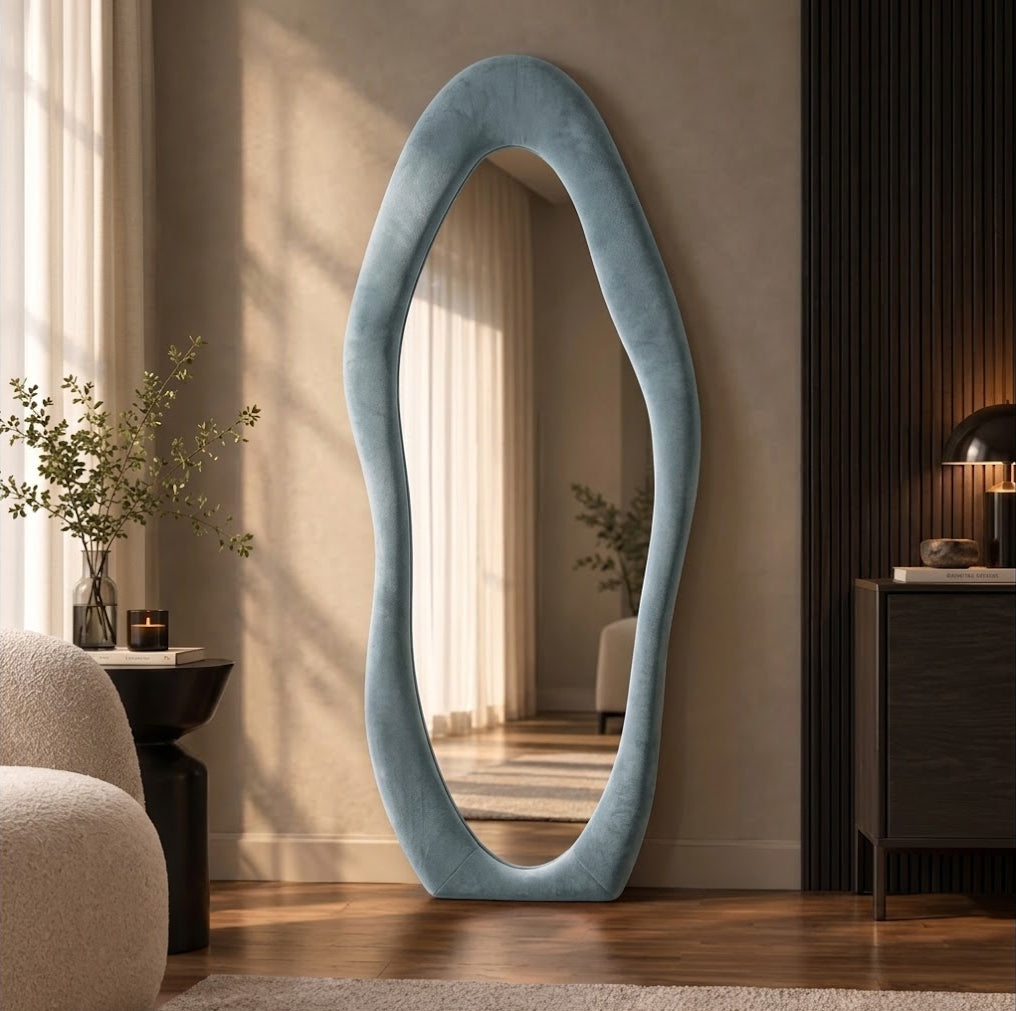 Blue-gray wavy flannel-framed floor mirror leaning beside a wood-slat wall, reflecting window curtains.