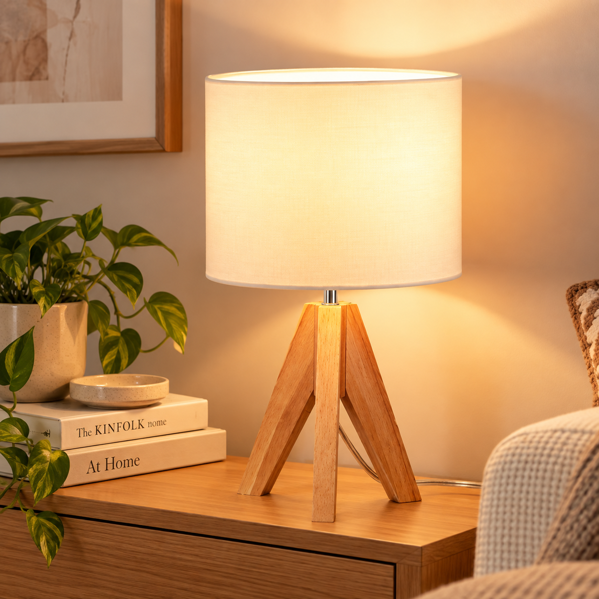 Natural wood tripod table lamp with warm glowing cream cylindrical shade on a wood side table beside an armchair, books, and a plant by HavvakHome.
